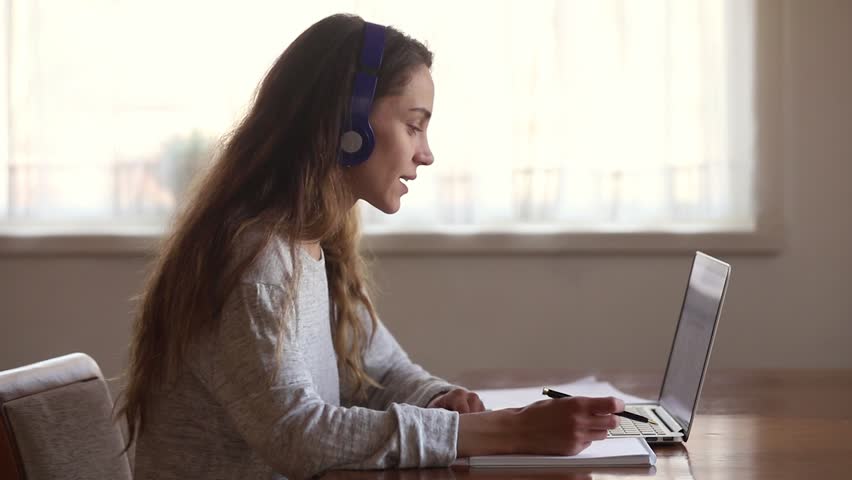 Serious girl student wear headphone study online with internet teacher learn language talk looking at laptop, focused young woman make video call tutoring write notes, teaching concept Royalty-Free Stock Footage #1029331781