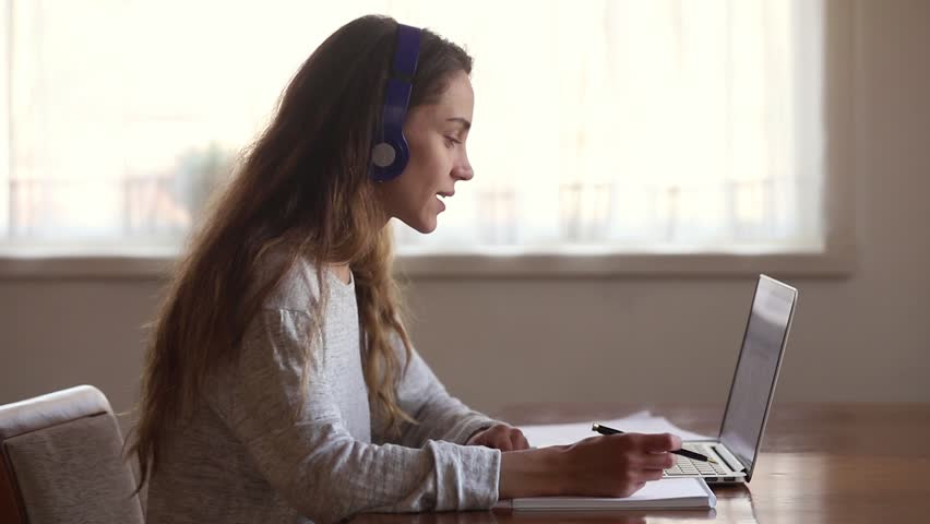 Serious girl student wear headphone study online with internet teacher learn language talk looking at laptop, focused young woman make video call tutoring write notes, teaching concept | Shutterstock HD Video #1029331781