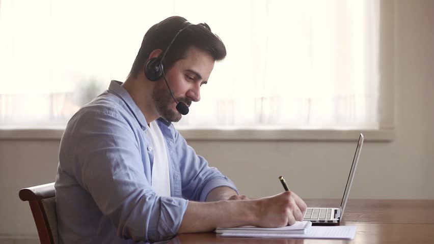 Focused business man interpreter teacher wearing headset looking at laptop screen making notes, male online tutor teaching student learning in internet video calling on computer writing down Royalty-Free Stock Footage #1029331784