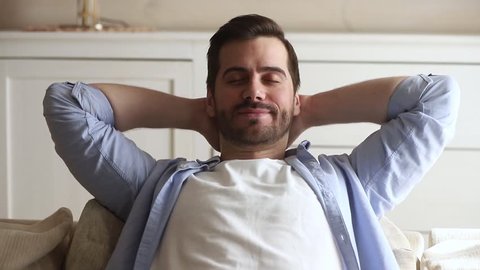 Calm happy young man relaxing with eyes closed on comfortable couch holding hands behind head breathing fresh air, lazy serene millennial guy chilling on sofa enjoy stress free peaceful day at home