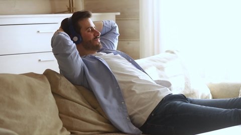 Calm young man wearing wireless headphones chilling listening to music sit on sofa, happy relaxed millennial guy enjoying lounge sound with eyes closed being in peaceful mood feeling no stress free