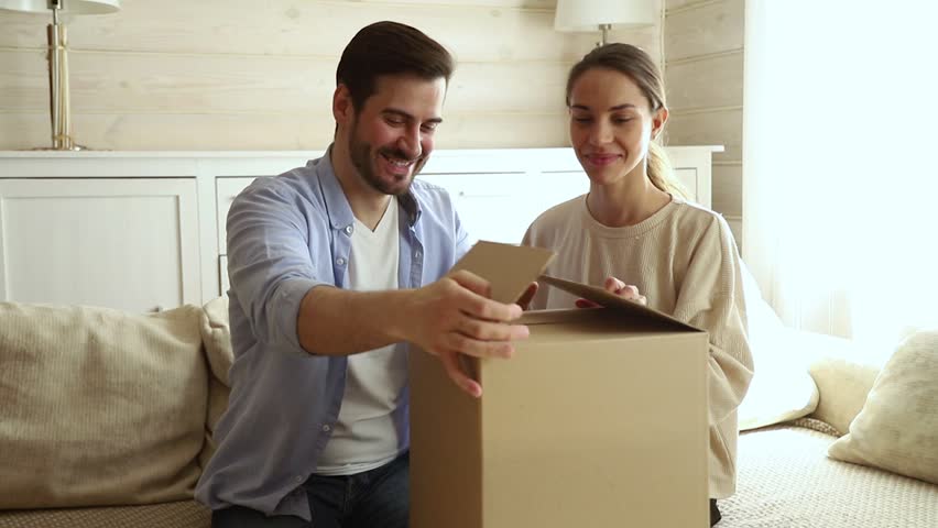 Happy excited couple customers open cardboard box together sit on sofa at home, young family consumers unpack good parcel looking inside receive surprising great purchase delivered by postal shipping | Shutterstock HD Video #1029331829