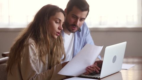 Serious young couple holding papers pay domestic bills online on laptop checking bank account reading document at home, millennial family planning budget discussing money finances expenses