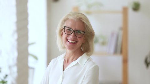 Head shot of fifty years woman wearing glasses pose in office room looking at camera laughing feels happy, independent elderly company owner, coach or leader successful businesswoman portrait concept