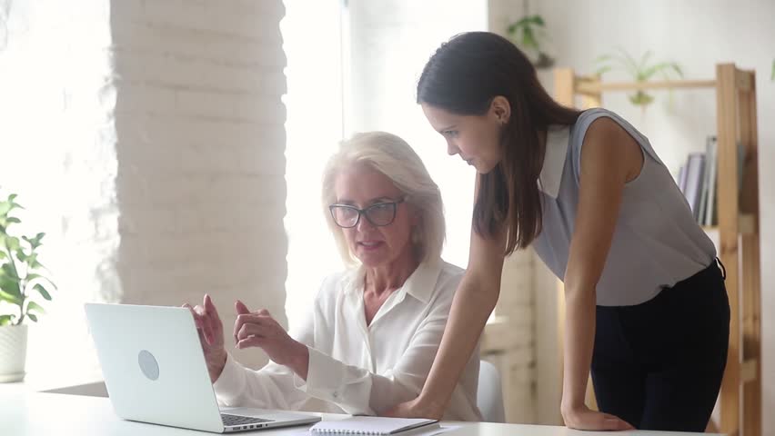 Elderly businesswoman manager ceo in glasses sit at desk typing on computer showing explaining program app to intern, do online task together, helping apprentice teach new employee, mentoring concept