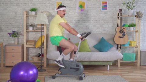 Funny energetic man from the 80s with a mustache engaged at home on a exercise bike slow mo