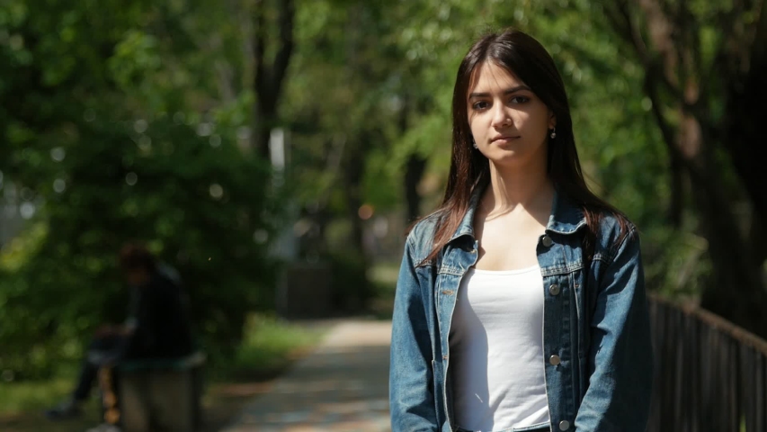 young caucasian woman with dark hair and jeans jacket standing in the park in a sunny day