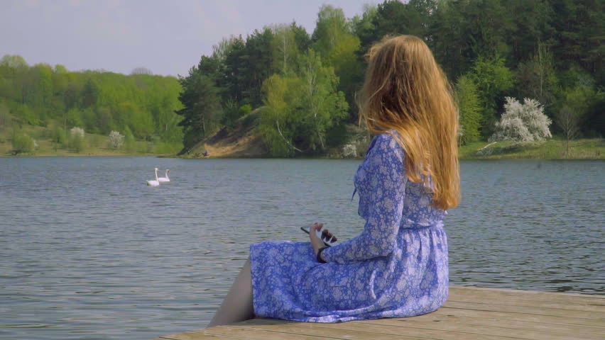 beautiful red-haired girl sitting on the pier by the lake. girl in blue dress looking at floating swans. slow motion shot.