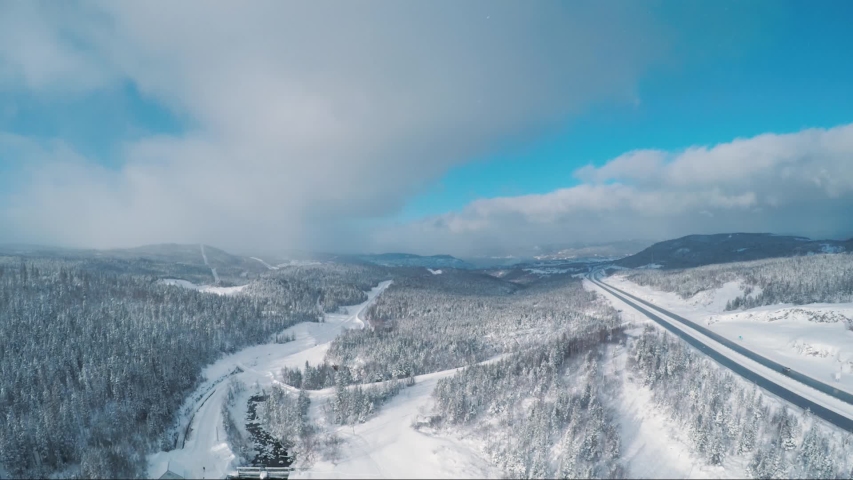 Aerial view shows cars driving down the highway in the winter. See snow falling in the sky and see white snow on the trees. This is Newfoundland, Canada. It is a sunny winter day.