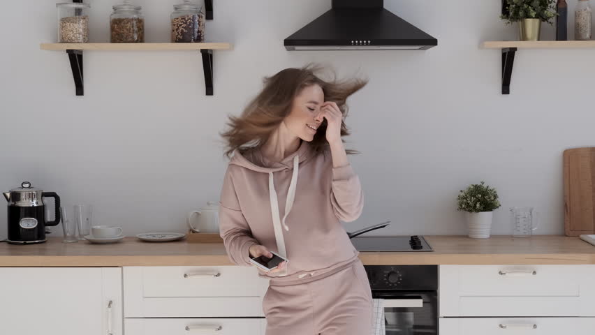 Energetic dancing of a happy girl. Pretty young woman dancing in kitchen and listening music. Beautiful woman listening to music on smartphone. Young female dancing and using cellphone Royalty-Free Stock Footage #1029350660