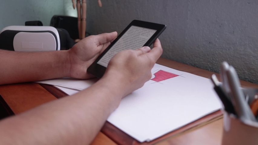 Side view. Male hand reading and turning pages on an ebook reader. Ebook reader on a wooden desk. Vr headset on background. 3d experience with a vr headset