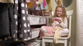 Beautiful little girl sitting in the home wardrobe uses the phone, watching a video or playing games. On the girl's lap are things