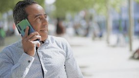 Man talking by cell phone and walking on street. Cheerful smiling Hispanic man having pleasant phone conversation outdoor. Connection concept