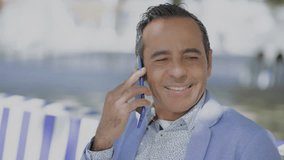 Happy mature man talking by cell phone outdoor. Smiling handsome middle aged man in suit jacket sitting on bench and having pleasant phone conversation. Connection concept