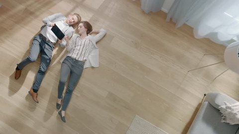 Young Couple are Lying on a Wooden Flooring in an Apartment. They are Using a Tablet Computer. Cozy Living Room with Modern Interior, Grey Sofa and Wooden Parquet. Top View Camera Footage.