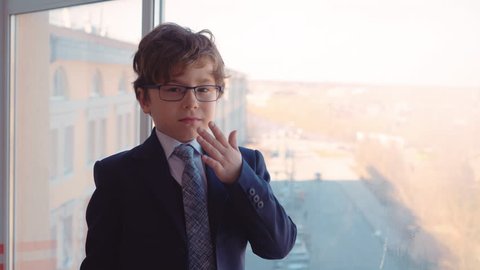 Cute Boy Wear Businessman Suit Look Office Window. Nice Caucasian Child in Glasses Put on Hat Adult Life Parody. Serious Kid Business Strategy Skyscraper Background Occupation Concept Closeup 4K