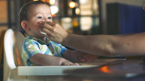 Mom wipes the baby boy's mouth and hands with a napkin. Cute kid eating his lunch. The adorable child laughs and claps his hands. The concept of the rules of etiquette, cleanliness while eating.
