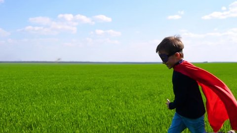 A happy joyful child in a superhero costume, a red cloak and a mask, runs across a green field and laughsの動画素材