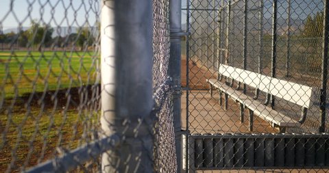 Slow pan across a chain link fence and an empty baseball field bench in the dugout of a local park at sunrise.