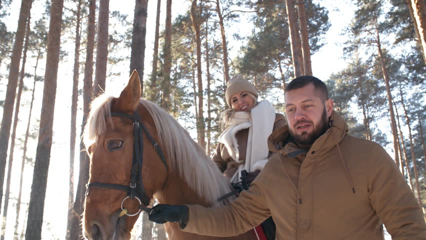 Low angled shot of woman riding horse and speaking with man while he holding leading reins and walking in forest on sunny winter day; sunlight shining at camera