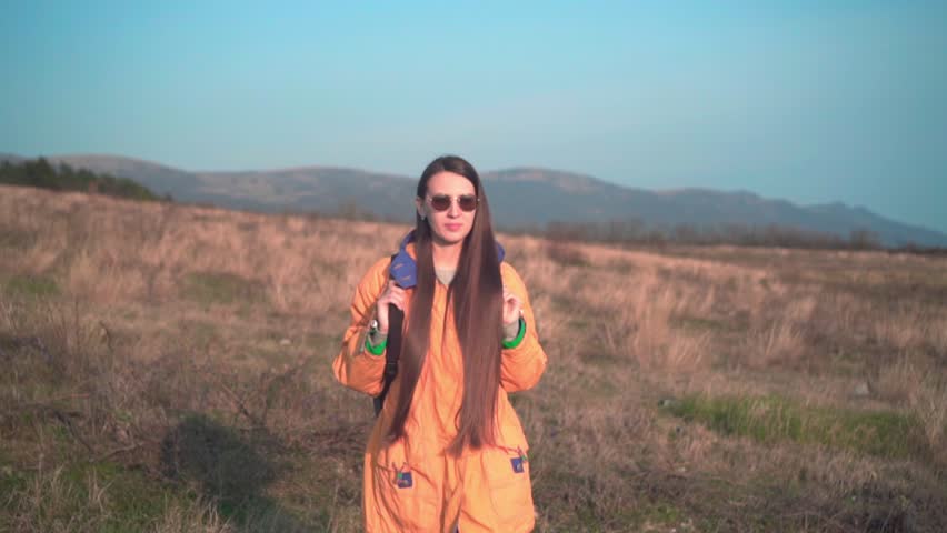 A young girl with long dark hair in a yellow jacket, a leather cowboy hat and glasses walks on the yellow grass, looks into the distance. Background mountains, sky.