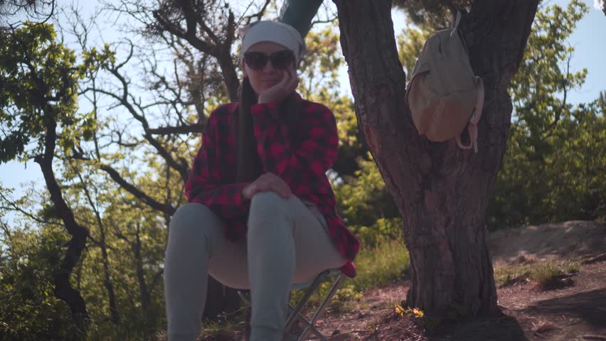 A girl with dark hair in a black and red shirt, a bandana and glasses sits in the forest, looking around, a tourist backpack hangs on a tree. Background sky, forest.