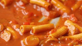 Close video of rigatoni pasta and slices of sausage in a tomato sauce being stirred with a wood spoon in a pan while heating illuminated with natural lighting.