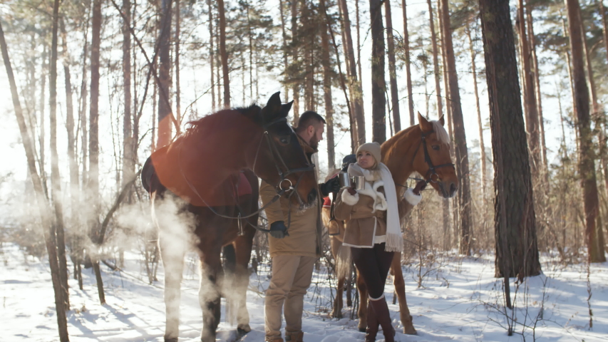 Romantic couple toasting with metal cups and drinking hot tea while standing with horses in forest on sunny winter day