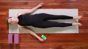 Top view of young woman doing Yoga exercise lying down in Shavasana Pose meditating with Smartphone.