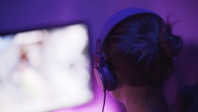 A shot of a female pro gamer from back left-side playing an online video game. Dark room with with purple and blue lights makes a good gaming mood.