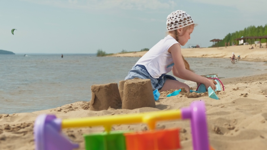 A preschool girl with blond hair plays on the beach by the river, builds a sand castle using a bucket, shovels and figures. Sunny summer day