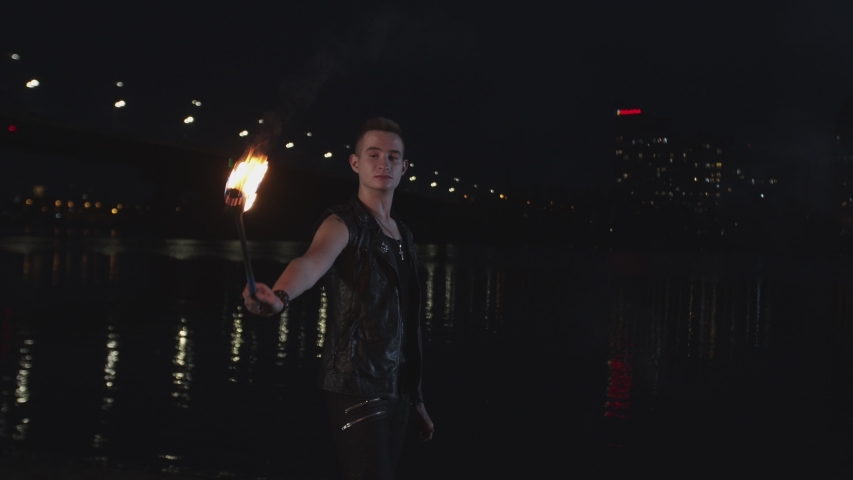 Young artist performing fire show oudoor at night time standing on river bank. Stylish man in leather clothes exhaling powerful fiery jet over night city lights reflected in dark water in background.