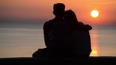 Silhouette of couple in love at sea in golden and beautiful sunset. Young and romantic couple enjoying.  Boy and girl on holidays on romantic summer evening. Happy couple talking and hugging on beach