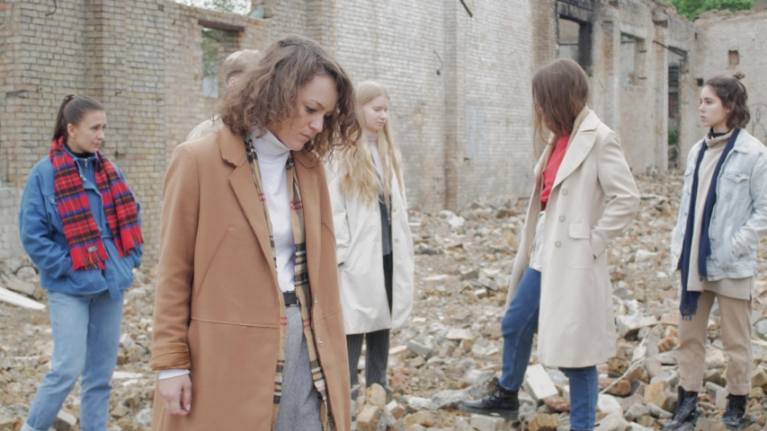 Curly woman in a coat performs a dance among a group of young people in the ruins. The youth makes a theatrical sketch against the background of a collapsed building of bricks Royalty-Free Stock Footage #1029449513