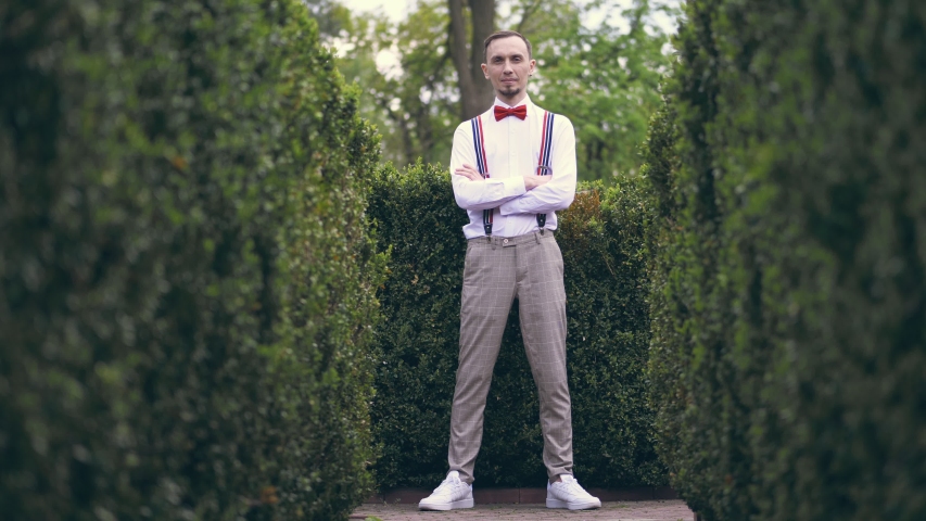 among the beautifully cropped bushes in the park, a young man in a white shirt, trousers, stylish suspenders and bow tie stands and smiles a little.