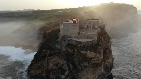 Aerial view of lighthouse on a cliff with a fortress on the coast of the Atlantic ocean  in Nazare, Portugal,  4k