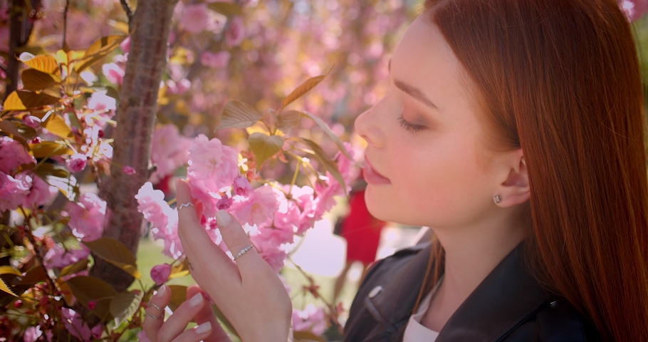 Close-up portrait of pretty ginger girl smelling the pink flowers tenderly in park.