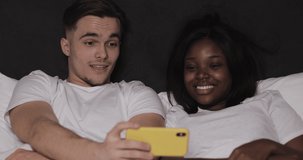 Happy multi-ethnic couple watching video together on the yellow smartphone lying in bed at night. They laughing and having fun. Relationships, family, social network, a good pastime concept.