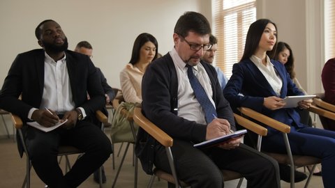 Diverse group of multinational business people taking part in corporate meeting. Bored african american man yawning while other members of seminar attentively listening speech and writing notes