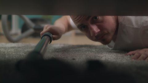 Young man cleaning a floor under a bed lying on the floor using vacuum cleaner