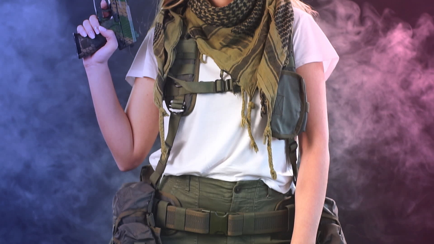 Woman in protective armour and headphones holds gun in hands, posing over dark smoky background. Army, dedication, weapon, technology.