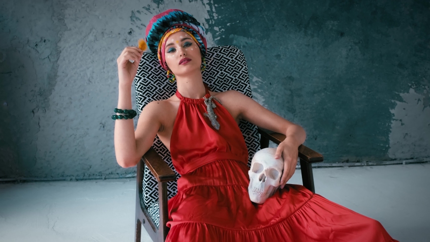 the model in the Eastern dress sits relaxed in a chair against the wall and holds an orange bird feather. camera