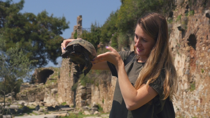 Young tourist woman in green t-shirt holding in hands old tortoise with ancient greek temple ruins on background.