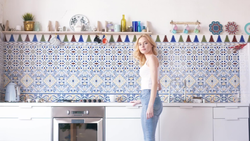 Close-up of beautiful young woman with blond hair in white t-shirt and blue jeans making a tea or coffee at the kitchen. Action. Relaxing at home