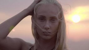 Close up of Attractive blonde woman posing at sunset, on top of a mountain, wind in her hair, in the late afternoon. Beauty shot golden hour 4 K in slow motion with blur. She is looking at camera.