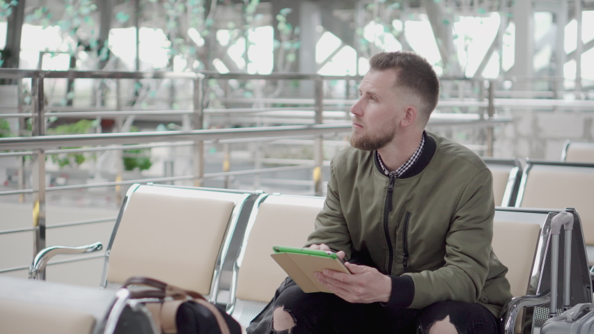 Young male traveler is putting tablet in his backpack, checking time on watch, sitting in waiting room of terminal. He is sitting on bench in departure lounge of airport alone