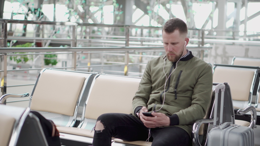 Handsome young male passenger is enjoying music through headphone in departure lounge of airport. He is outputting headset and listening announcement