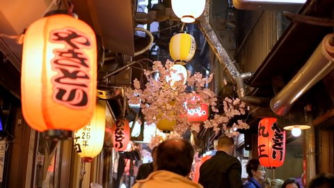 Tokyo, Japan - April 4, 2019: Memory lane piss alley with cherry blossom decorations and many people walking by izakaya bars in spring in Shinjuku area of city at night in slow motion