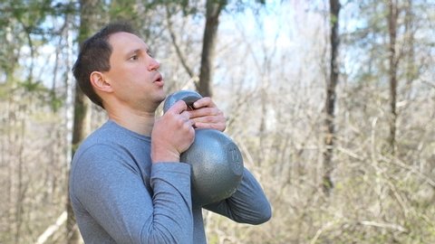 Young fit man exercising with heavy kettlebell in outdoors outside park holding weight lifting doing over the shoulder exercise in slow motion