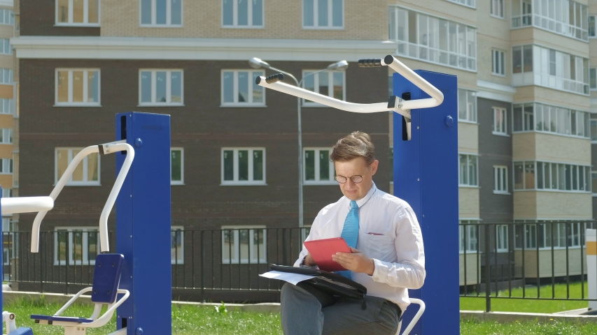 A businessman sits on a sports simulator outdoors, studies contracts and uses a mobile phone. Remote work. The concept of a healthy and sporty lifestyle